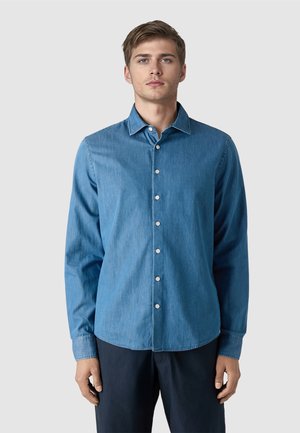 Junger Mann mit kurzen braunen Haaren, der ein langärmliges blaues Hemd mit Knöpfen und dunkle Hosen trägt, vor einem schlichten hellgrauen Hintergrund stehend.