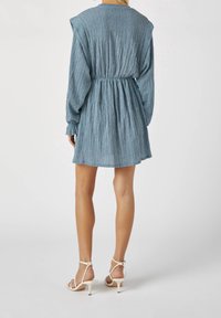 Mujer vestida con un mini vestido azul de manga larga texturizado y cintura fruncida, de pie, de espaldas, con sandalias de tacón alto blancas y de tiras.
