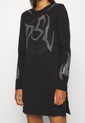 Robe longue noire à manches longues avec des motifs géométriques abstraits argentés sur la poitrine et les manches, portée par une personne debout, les mains détendues.