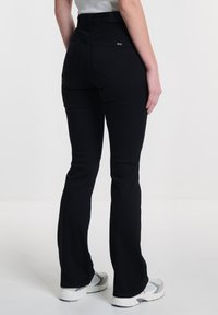 Jeans de campana negros con una cintura de altura media, costuras sutiles, dos bolsillos traseros y una textura suave. Combinados con zapatillas deportivas grises.