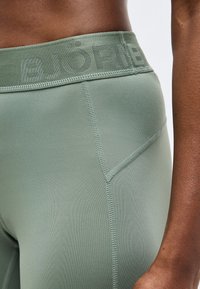 Κοντινή λήψη ενός ατόμου που φορά ανοιχτό πράσινο κολάν με εμφανείς ραφές και ζώνη μέσης με λογότυπο που γράφει «BJÖRN».