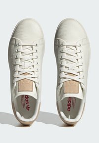 Hvide lærredssneakers med tekstureret stof, flade snøreb�ånd og lysebrune accenter. Har et logo-patch på tungen og en rød trykt indersål.