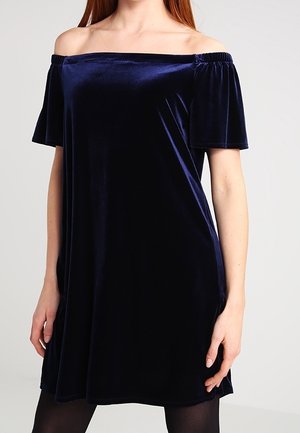 Femme portant une courte robe en velours bleu marine à épaules dénudées avec manches courtes et des collants noirs, debout devant un fond uni.