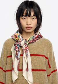 Pañuelo de seda con vibrantes patrones florales y de cachemira en tonos de rojo, verde y azul, atado alrededor del cuello sobre un suéter beige con rayas rojas.