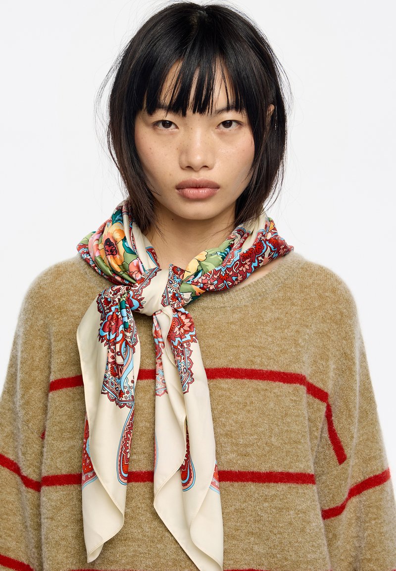 Pañuelo de seda con vibrantes patrones florales y de cachemira en tonos de rojo, verde y azul, atado alrededor del cuello sobre un suéter beige con rayas rojas.