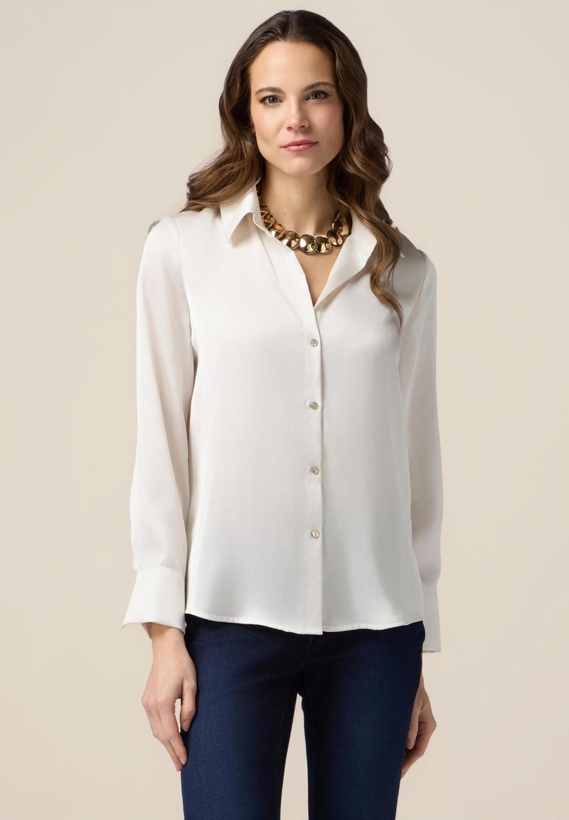 Blusa bianca a maniche lunghe realizzata in tessuto morbido, con colletto, chiusura a bottoni nella parte anteriore e maniche con polsini, abbinata a jeans scuri.