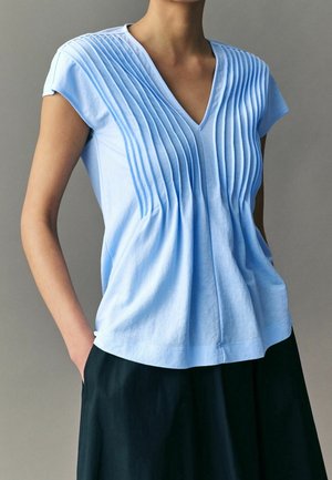 Personne portant un chemisier bleu clair à manches courtes avec des plis verticaux et un pantalon noir, debout avec une main dans la poche devant un fond neutre.