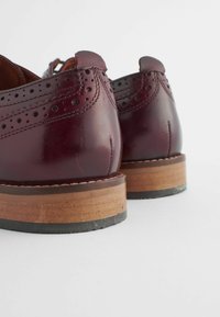 Chaussures en cuir bordeaux avec des détails perforés, bout rond et talons en bois empilés. Texture lisse avec une finition brillante.