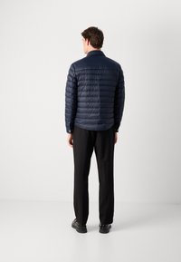 Mornarica puhovka s kvadratnim teksturo, stojnim ovratnikom in nazaj yoke, v kombinaciji s črnimi hlačami in črnimi čevlji.