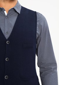 Námořnická pletená vesta se šesti knoflíky a náprsní kapsou, navrstvená přes vzorovanou modro-bílou košili. Texturovaná látka, přiléhavý střih.