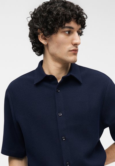 Νέος άνδρας με καστανά σγουρά μαλλιά, φορώντας μπλε σκούρο κοντομάνικο πουκάμισο με κουμπιά, κοιτάζει προς τα πλάγια μπροστά σε απλό φόντο.