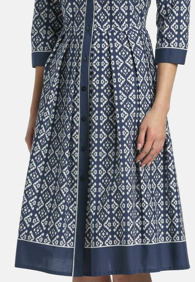 Blau-weiß gemustertes Hemdkleid mit Dreiviertelärmeln und einem einfarbigen blauen Saum, abgebildet von der Mitte des Oberkörpers bis zu den Knien.