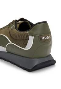 Olivově zelené a bílé tenisky s texturovanou látkou, černou podrážkou a logem "HUGO" na patě, zobrazené z pohledu zadního úhlu.