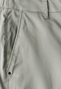 Tissu vert clair avec une texture douce, comportant une poche avant et des coutures visibles. Comprend un bouton noir pour la fermeture.