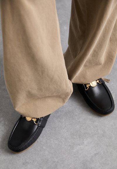 Mustad nahast loafers kuldse ahelaga aksessuaariga ja õmblustehnikaga, millega on paaritatud heledast tanist püksid, uduselt hall taust.