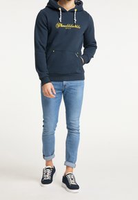 Marinblå huvtröja med gul text och vita dragsnören, framfickan i kängurstil, kombinerad med ljusblå jeans och marinblå sneakers. Casual passform.