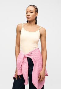 Νέος με πλεγμένα μαλλιά, που φοράει μπεζ αμάνικο τοπ και σκούρο παντελόνι, με μια ροζ ριγέ πουκαμίσα δεμένη γύρω από τη μέση του.