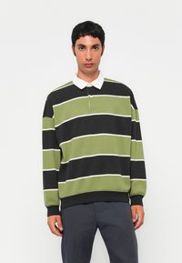 Ung mann kledd i svart og olivengrønnstripet langermet poloshirt med hvit krage og mørk grå bukser, stående mot hvit bakgrunn.