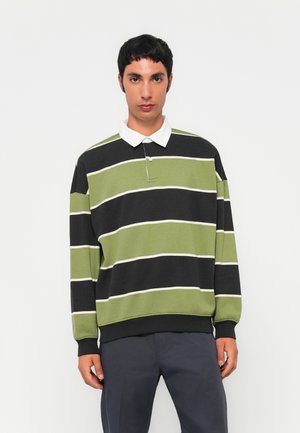 Ung mann kledd i svart og olivengrønnstripet langermet poloshirt med hvit krage og mørk grå bukser, stående mot hvit bakgrunn.