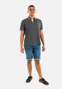 Camisa polo gris con cuello y ribete blanco, combinada con pantalones cortos de denim enrollados. Lleva zapatillas oscuras con acentos naranjas.