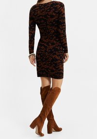 Femme de dos portant une robe longue à manches longues noir et marron à motifs, ainsi que des bottes en daim marron à talons hauts arrivant au genou.