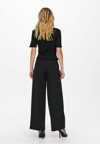 Black short-sleeve top with a scalloped hem, paired with wide-leg black pants. Smooth fabric with a relaxed fit; worn with black heels.