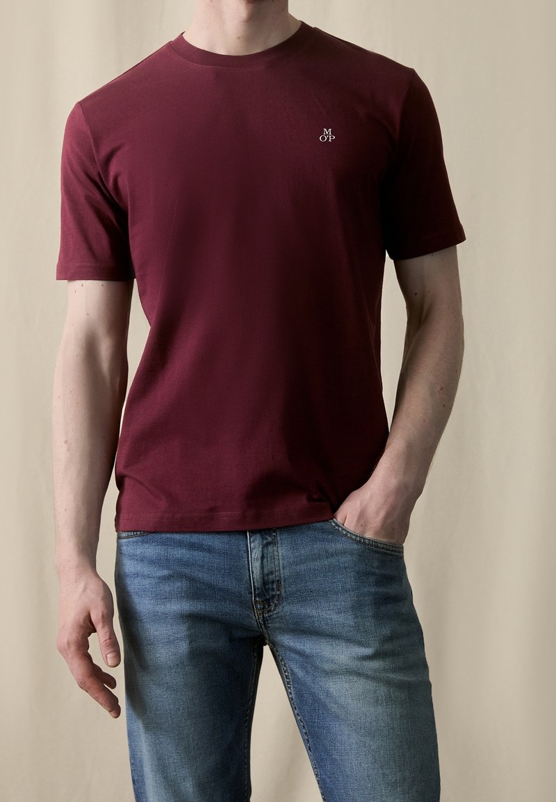 Homme portant un t-shirt uni bordeaux à manches courtes avec un petit logo blanc sur la poitrine, associé à un jean bleu, la main dans la poche.