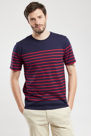 Mand med kort brunt hår og skæg iført marineblå t-shirt med røde vandrette striber og beige bukser, poserer med hænderne i lommerne.