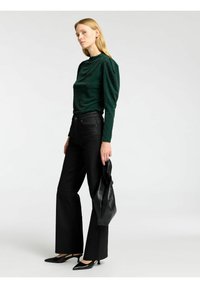 Mörkgrön, strukturerad långärmad topp med hög krage; kombinerad med svarta, utsvängda jeans och svarta klackskor. En svart handväska hålls i handen.