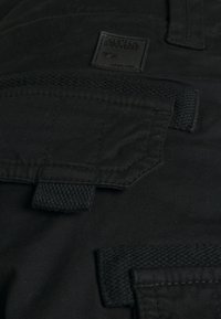 Gros plan sur un tissu noir avec des poches cousues et des boucles tissées, avec une petite étiquette portant le nom de la marque et le logo.
