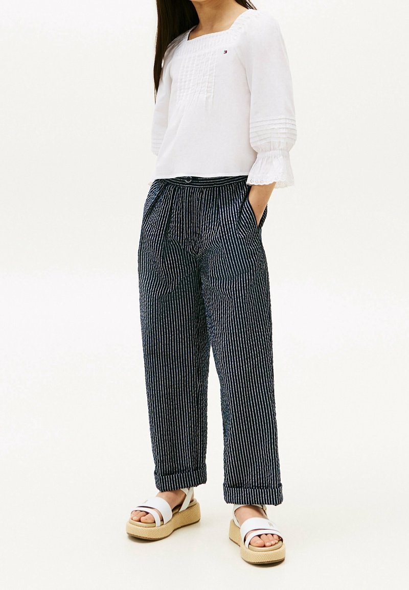 Femme portant un chemisier blanc à manches trois-quarts, un pantalon large à fines rayures bleu marine, et des sandales compensées blanches, debout, les mains dans les poches.