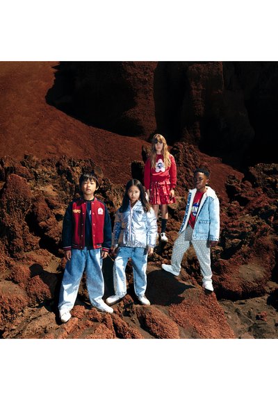 Quatre enfants se tiennent sur un terrain rocheux. Leurs tenues comprennent une veste universitaire rouge, une doudoune argentée, une veste en jean et un pantalon à motifs.