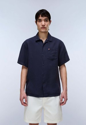 Junger Mann trägt ein marineblaues kurzärmeliges Hemd mit Knöpfen und weiße knielange Shorts, steht vor einem schlichten hellgrauen Hintergrund.