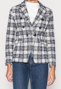 Blazer de doble botonadura en un patrón de cuadros azul marino y blanco, con botones grandes, un corte entallado y dos bolsillos delanteros.