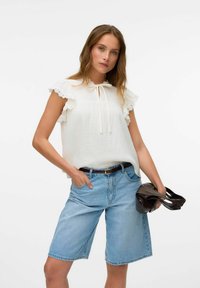 Vit strukturerad blus med volangärmar och knytkrage, kombinerad med ljusblå denimshorts. Modellen håller en svart väska.