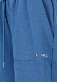Siniset puuvillasekoiteshortsit kiristysnyörillä, joissa on tasku ja valkoinen "LES' ARCS"-logo. Pehmeä pinta, rento istuvuus.