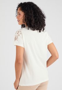 Camiseta de color claro con mangas cortas y un intrincado detalle de encaje en los hombros,fabricada en una tela suave con un corte relajado.