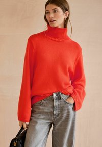 Jasnoczerwony sweter z golfem wykonany z dzianiny, z ribbowanym kołnierzykiem, w połączeniu z szarymi dżinsami o wysokim stanie. Modelka trzyma małą czarną torebkę.