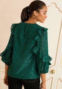 Blusa teal con texture e maniche a volant, apertura a chiave sul retro e tessuto leggermente trasparente. Abbinata a jeans neri per un contrasto.