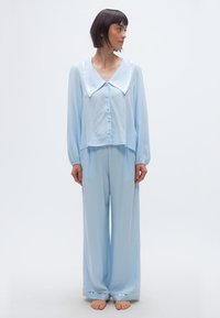 Γυναίκα με κοντά σκούρα μαλλιά που φοράει ανοιχτό μπλε φαρδιά πυτζάμα με μακριά μανίκια και φαρδιά παντελόνια, όρθια ξυπόλητη σε λευκό φόντο.