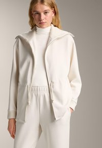 Biela zipsová bunda s rebrovaným golierom a manžetami, s nastaviteľným pásom a bočnými vreckami, kombinovaná s vyhovujúcim rolákom.