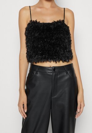 Frau trägt ein schwarzes, pelziges Crop-Top mit Spaghettiträgern und hoch taillierte schwarze Lederhose, gezeigt von Hals bis Hüfte.