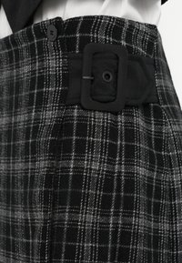 Čierne a sivé kárované nohavice so širokým opaskom, ktorý má štvorcovú pracku a vertikálnu textúru. Na opasku je viditeľné zapínanie na gombíky.