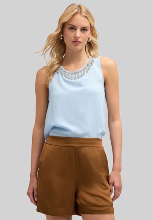 Top senza maniche azzurro chiaro con decorazioni a perline e frange sul collo, abbinato a pantaloni corti di raso marroni. Tessuto dalla texture liscia.