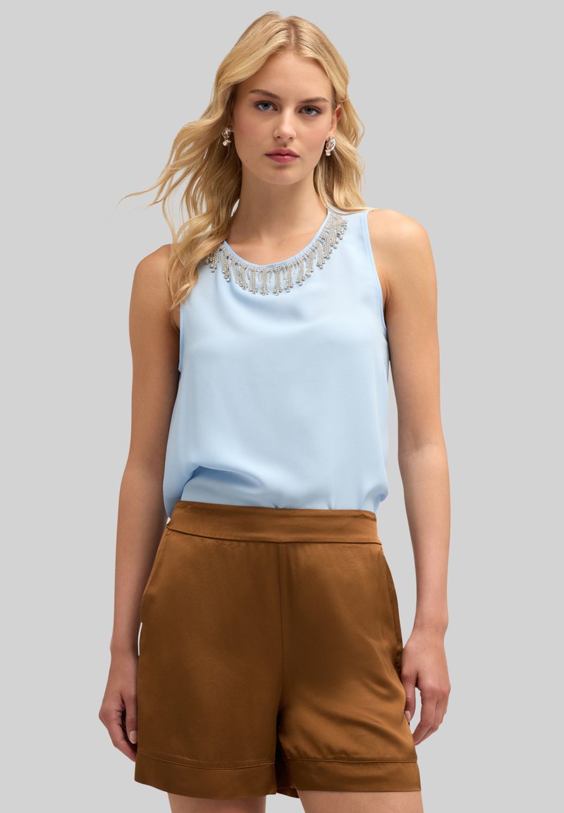 Ljust blå ärmlös topp med dekorativa pärl- och fransdetaljer vid halslinningen, matchad med bruna satängshorts. Slät tygkänsla.