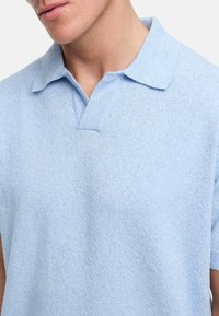 Lys blå kortermet polo-skjorte laget av myk, strukturert stoff. Har klassisk krage og en subtil lomme på venstre bryst.