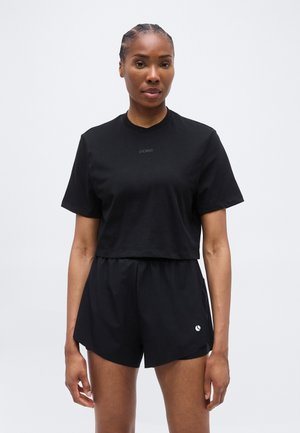 Žena s pletenicama, odjevena u crnu kratku majicu i crne sportske kratke hlače, stoji ispred jednostavne svijetle pozadine.