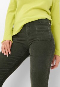 Pantalon en velours côtelé vert olive avec une surface texturée, doté de poches avant et arrière. Associé à un pull vert lime vif.