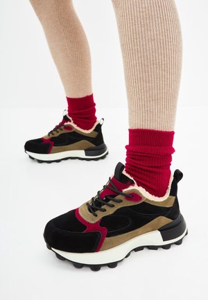 Black, olive, and red sneakers with thick white soles worn with red socks and beige ribbed leggings against a white background.