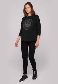 Sudadera negra con cuello redondo, mangas tres cuartos y un estampado gráfico. Combinada con jeans ajustados negros y zapatillas deportivas negras.